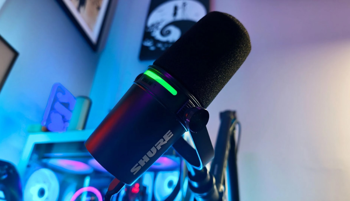 Динамічний мікрофон Shure MV7+ чорного кольору на пантографі з увімкненою зеленою світлодіодною LED-смугою та білим логотипом SHURE на корпусі в неоновій кімнаті стрімера