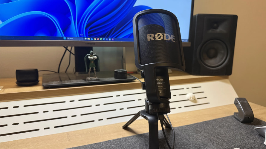 Мікрофон RODE NT-USB+ на столі перед монітором комп'ютера з синім внутрішнім підсвічуванням капсюля та написом REVOLUTION PREAMP